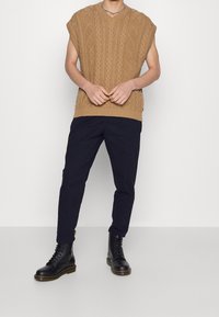 Maglione senza maniche di colore marrone chiaro, lavorato a maglia con scollo a V, caratterizzato da un motivo a diamante testurizzato; abbinato a pantaloni blu navy e stivali neri con lacci.