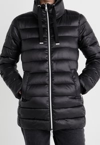 Černá péřová bunda s prošívaným vzorem, vysokým límcem, předním zipem a dvojitými stahovacími šňůrkami. Má boční kapsy a hladkou texturu.