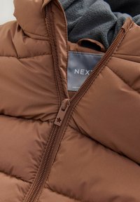 Giubbotto marrone imbottito con design trapuntato, dotato di chiusura con zip e un morbido rivestimento in pile grigio al collo. Etichetta del marchio visibile all'interno.