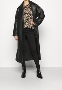 Cappotto lungo in finta pelle nera sopra una maglietta con stampa leopardata, abbinato a pantaloni neri e stivaletti con tacco neri. Texture liscia e vestibilità sagomata.