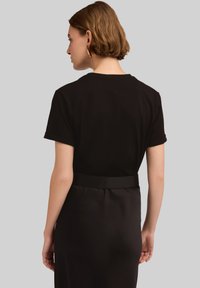 Camiseta de algodón negra con mangas cortas, vista desde atrás. Combinada con una falda de tela negra suave con cinturilla.