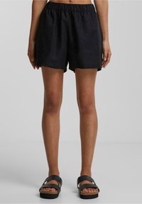 Personne portant un short noir à taille élastique et des sandales noires à semelles en liège, se tenant contre un fond gris clair uni.