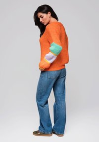 Helles orange gestricktes Sweatshirt mit mehrfarbigen Rippärmeln, kombiniert mit lockeren blauen Jeans und braunen Schuhen.