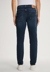 Jeans en denim bleu foncé avec une coupe droite, présentant une étiquette en cuir sur la ceinture arrière et deux poches plaquées avec un détail de couture.