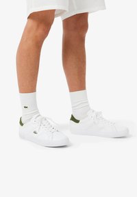 Sneakers bianche con accenti verdi sul tallone, tomaia in pelle testurizzata e dettagli laterali traforati. Abbinati a calzini bianchi alti.