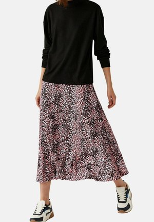 Žena v černém tričku s dlouhým rukávem, růžovo-černé vzorované midi sukni a bílých a námořnicky modrých teniskách, stojící před jednobarevným pozadím.