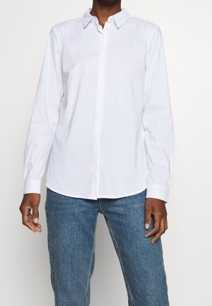 Personne portant une chemise blanche unie à manches longues boutonnée et un jean bleu, debout devant un fond uni clair.