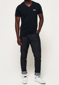 Marineblauwe v-hals t-shirt, korte mouwen, gedragen met donkere spijkerbroek en witte sneakers. Bevat logo op de borst en manchetten. Schoenen hebben veters en een minimalistisch ontwerp.