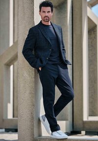 Blazer aus schwerem, strukturiertem Stoff über einem schwarzen Hemd, kombiniert mit dunkler Hose und weißen Sneakers, die an einem Betonsäule lehnt.