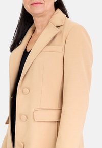 Abrigo texturizado beige con un ajuste entallado, que presenta un cuello con muesca, cierre de doble botón y bolsillos laterales.