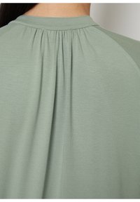 Camisa verde con detalles fruncidos en la parte posterior, que presenta una textura suave y un diseño clásico de cuello redondo. Tejido suave con un ajuste drapeado.