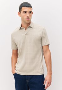 Hombre con pelo corto y oscuro, vistiendo una camiseta polo de manga corta beige con un patrón y vaqueros azul oscuro, de pie con una mano en el bolsillo.