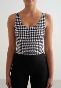 Juoda ir balta hounds-tooth rašto liemenėlė su apvaliu iškirpimu, derinama su aukštu liemeniu juodomis kelnaitėmis, kuriose yra subtilus prekės ženklas.