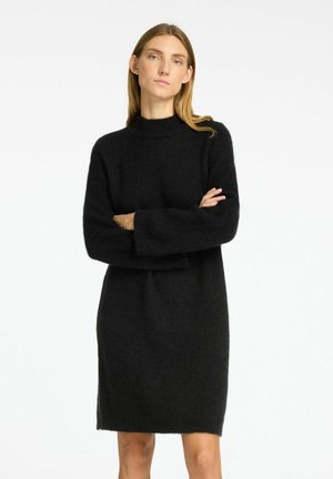 Černé pletené šaty s vysokým límcem, oversized rukávy a uvolněným střihem. Textura vypadá měkká a nadýchaná. Délka ke kolenům.