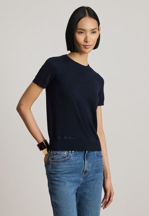 Donna con capelli neri e lisci che indossa una maglietta nera a maniche corte, jeans blu e un grande braccialetto al polso sinistro, in piedi con una mano in tasca.