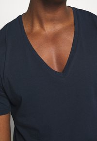 Primo piano di una persona che indossa una maglietta a V di colore blu navy scuro, mostrando la parte superiore del petto e la zona del collo con una tonalità di pelle medio-scura.