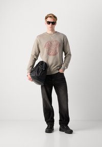 Grå hettegenser med rødt rundt logo, svarte jeans, svarte joggesko og solbriller. Bærer en svart snørebag.