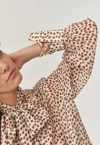 Femme en blouse beige à pois marron, avec un grand nœud au col et des manches bouffantes, portant un bracelet en or, posant avec une main derrière la tête.
