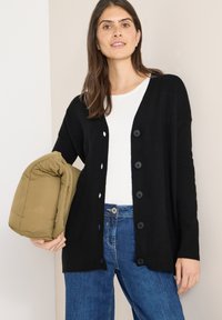 Černý cardigan se šesti knoflíky, žebrovanými manžetami a volným střihem, nošený přes bílou košili. V jedné ruce je držen zelený péřový kabát.