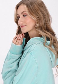 Una sudadera de color verde agua claro con puños acanalados y capucha con cordón. La tela parece suave con un ajuste cómodo y relajado.
