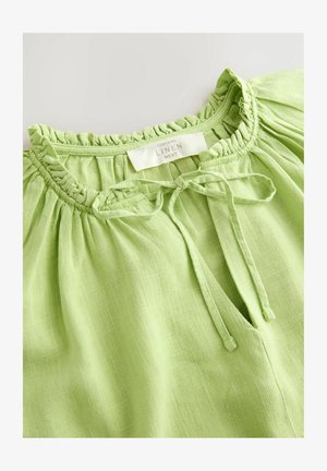 Linu blūze spilgti zaļā krāsā ar kroku izgriezumu un saitīti; izgatavota no teksturēta, gaisa materiāla ar nelielu pūku, lai nodrošinātu ērtu pieguļumu.