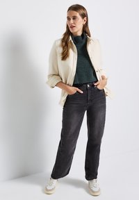 Crèmekleurig button-up shirt over een donkergroene trui, gecombineerd met zwarte rechte jeans en witte sneakers met beige accenten.