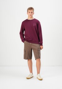 Mørkerød hettegenser med GAP-logo, beige cargoshorts med sidelommer, og hvite sneakers med beige såler. Uformell stil.