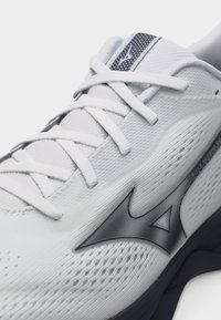 Λευκό αθλητικό παπούτσι με υφασμάτινο δίχτυ με υφή, λευκά κορδόνια και μαύρο λογότυπο Mizuno στο πλάι πάνω από μαύρη σόλα.