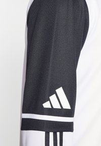 Adidas sportsjakke med en sort og hvid farveskeme, tekstureret stof og tre striber som accent samt et fremtrædende logo på ærmet.