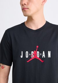 T-shirt en coton noir avec un logo Jumpman rouge en plein centre et "JORDAN" en texte blanc, avec un col rond et des manches courtes.