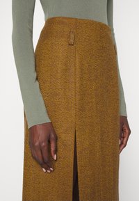 Brun kjol med fiskbensmönster och slits, parat med en figurnära grön långärmad topp. Texturerat tyg med skräddarsydd design.