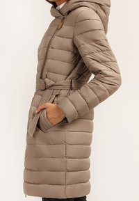 Žena v délce po kolena v taupe prošívaném kabátu s kapucí, opaskem a rukou v kapse, zobrazená ze strany na světlém pozadí.