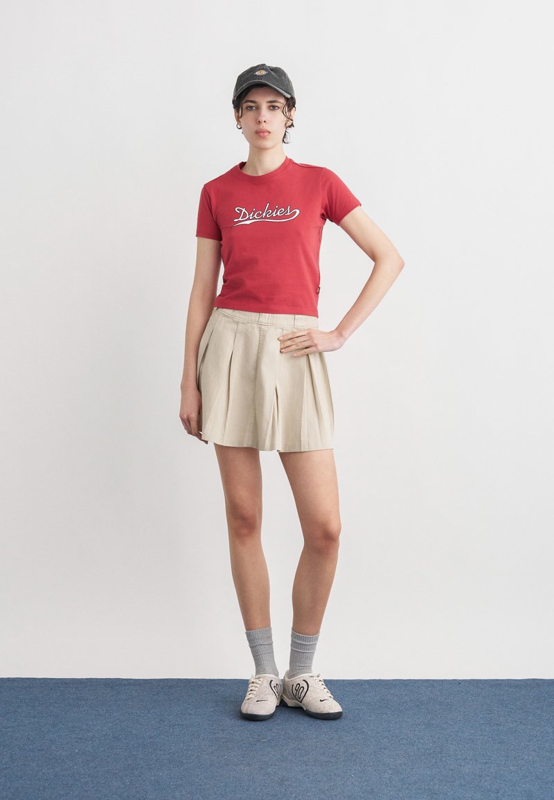 Giovane donna con berretto nero, maglietta rossa Dickies, gonna beige a pieghe, calzini grigi e scarpe da ginnastica bianche in piedi su un tappeto blu.