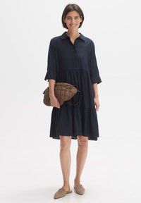 Dunkelmarineblaues Shirtkleid mit einem Kragen, Dreiviertelärmeln, gestuftem Saum und einer glatten Stofftextur. Hält eine braune gefütterte Tasche.