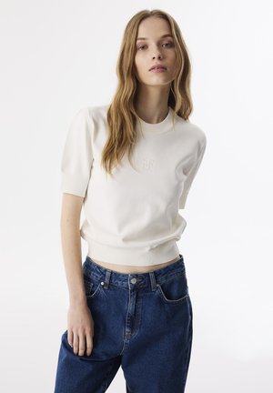 Maglione a maglia color crema con maniche corte e orlo a costine, caratterizzato da dettagli ricamati. Abbinato a jeans in denim blu a vita alta.