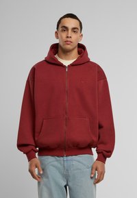 Red zip-up hoodie with a front pocket and adjustable hood. Features ribbed cuffs and hem with a subtle logo detail on the chest.