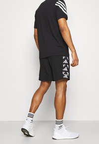 Hombre que lleva pantalones cortos negros de Adidas con detalles del logo en blanco, camiseta negra con rayas blancas en las mangas, calcetas blancas de Adidas y zapatillas blancas.