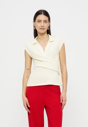 Femme aux longs cheveux foncés portant un haut cache-cœur crème sans manches et un pantalon rouge, debout devant un arrière-plan clair uni.