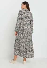 Langes Blumenkleid in Schwarz mit weißen Blüten, mit langen Ärmeln, einem gestuften Design und einem Kragen. Kombiniert mit hellen Sneakers.