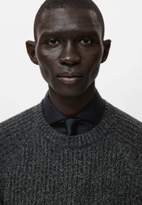 Maglione grigio lavorato a maglia con texture a coste, indossato sopra una camicia nera con cravatta, con scollo rotondo e primo piano del volto.