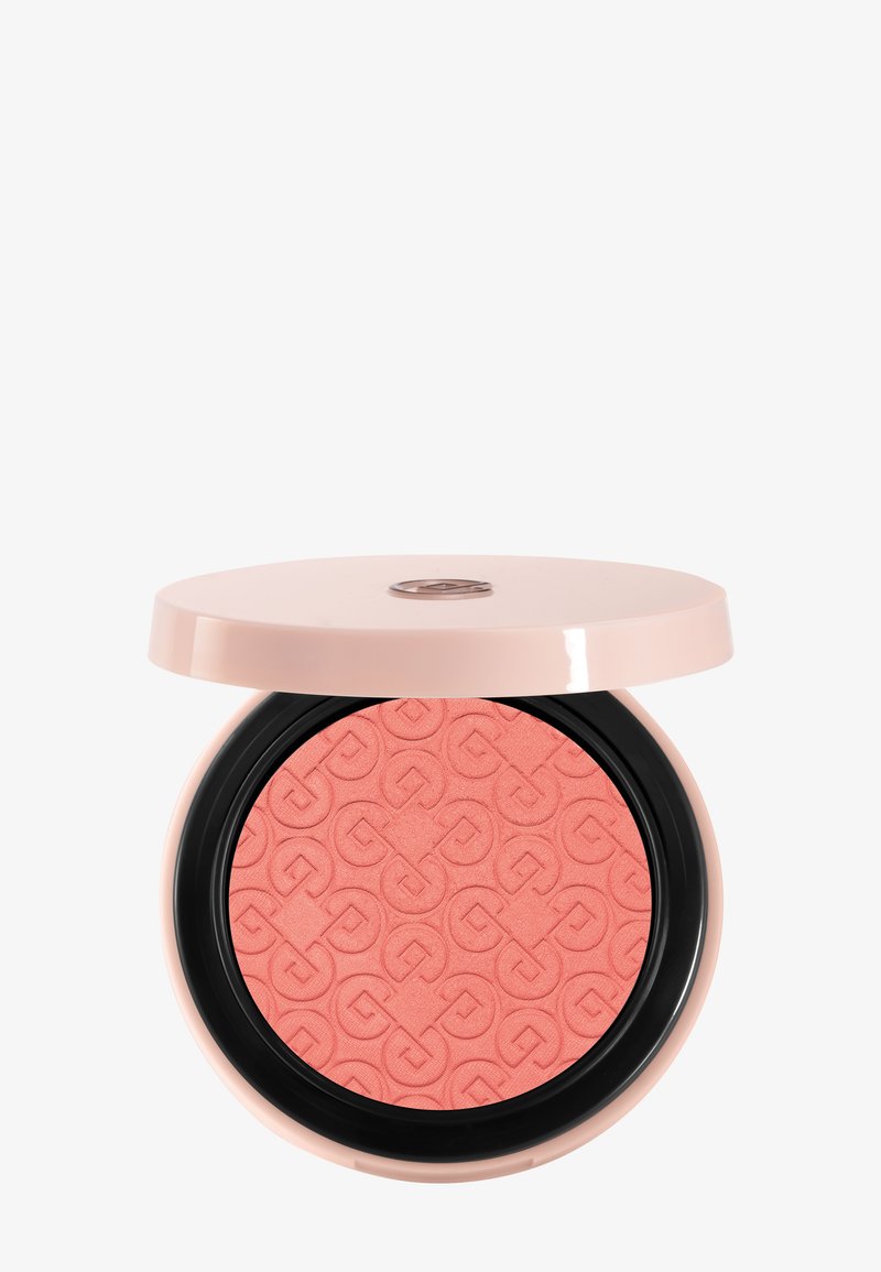 Blush corail compact rond avec un motif géométrique en relief, logé dans un boîtier rose pâle avec un anneau intérieur noir et un couvercle partiellement ouvert.