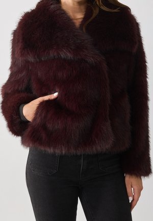Mujer que lleva una chaqueta corta de piel sintética burdeos oscuro con mangas largas, combinada con vaqueros negros de talle alto, mano en el bolsillo de la chaqueta.