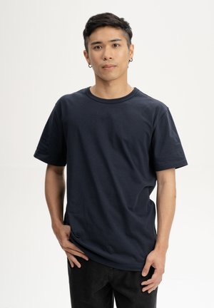 Jonge man met kort donker haar, gekleed in een effen donkerblauw T-shirt en zwarte broek, staand met één hand in de zak, neutrale achtergrond.