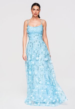 Femme portant une robe longue fleurie bleu clair avec de fines bretelles, debout devant un fond clair uni.