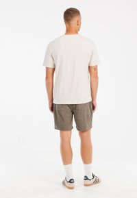 En lys beige kortærmet t-shirt parret med olivengrønne corduroy-shorts. Shortsene har to baglommer. Modellen står med ryggen til kameraet.