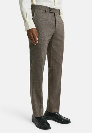 Uomo che indossa pantaloni a spina di pesce marroni, mokassini neri e una camicia a maniche lunghe color crema, in piedi contro uno sfondo bianco.