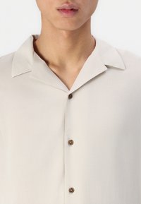 Light beige short-sleeve shirt with a collar, featuring brown buttons and a smooth, slightly shiny texture. Simple and modern design.