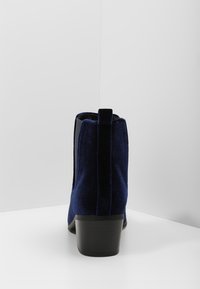 Botas de tobillo de terciopelo azul marino con un tacón bajo, paneles elásticos en los laterales y una pestaña en la parte trasera. Textura suave, diseño simplista.