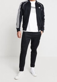 Svart jacka med vita ränder, matchad med svarta byxor och vita sneakers. Har en rund hals och en logotyp på bröstet.