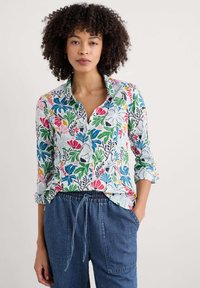 Chemise à boutons florale en tissu léger. Présente des imprimés colorés de feuilles et de fleurs, avec un col et des manches retroussées.